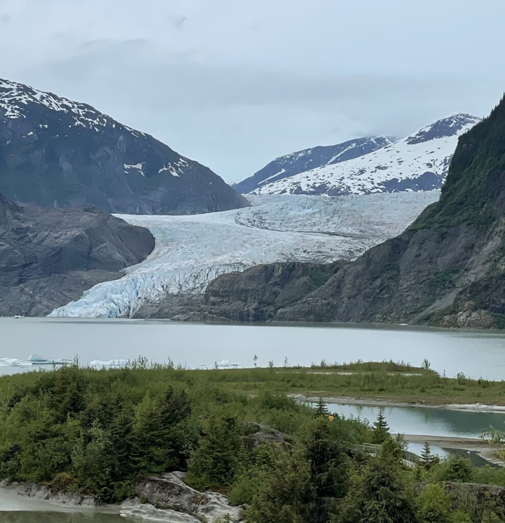 The blue-white face of Mendenhall Glacier between forested mountains, above the waters of Mendenhall Lake