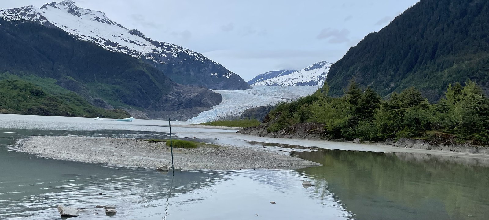 Mendenhall Glacier flowing down between snowy mountains to Mendenhall Lake in Juneau, Alaska, with a gravel shore in the foreground